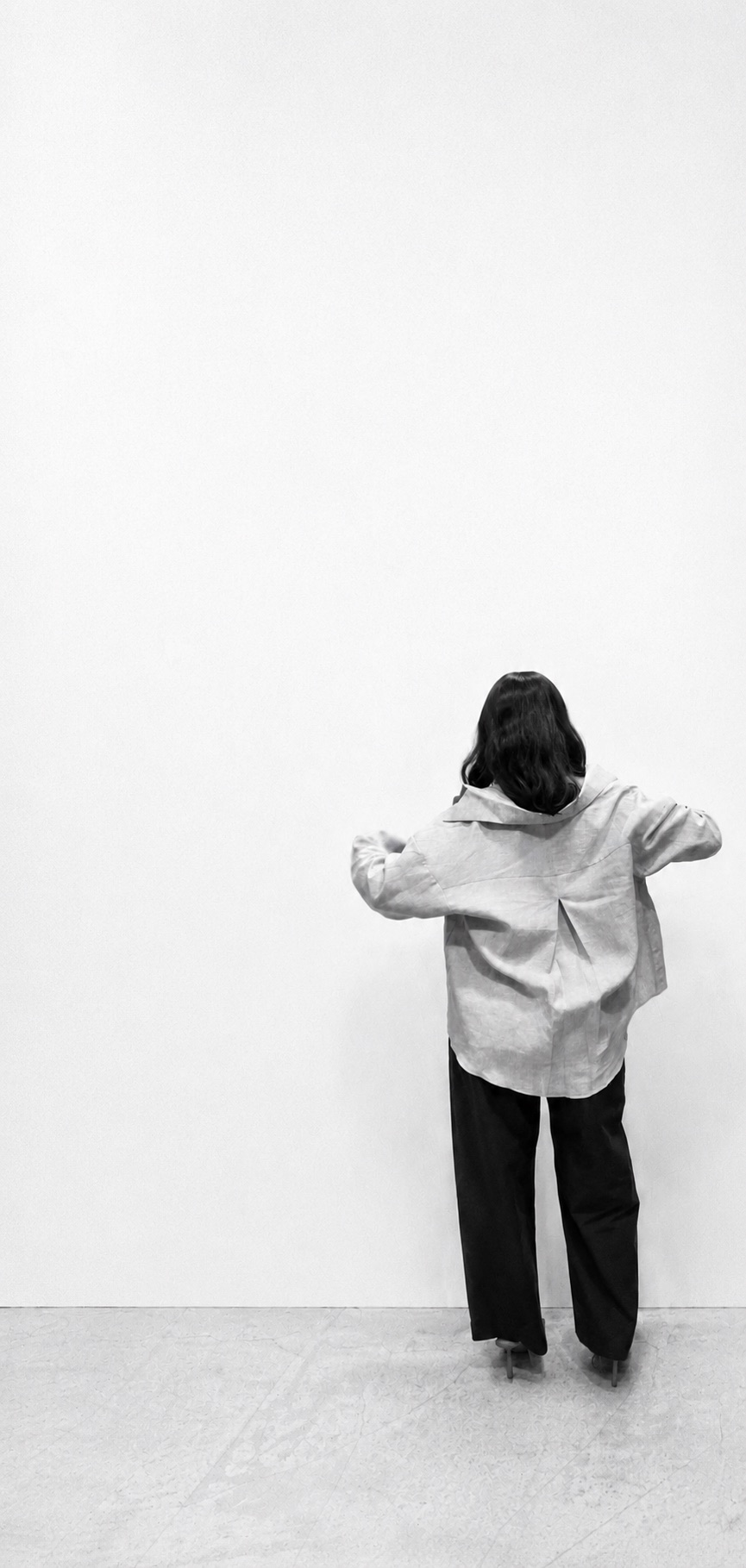 A woman stands facing a bare wall, adjusting the cuff of an oversized linen shirt.